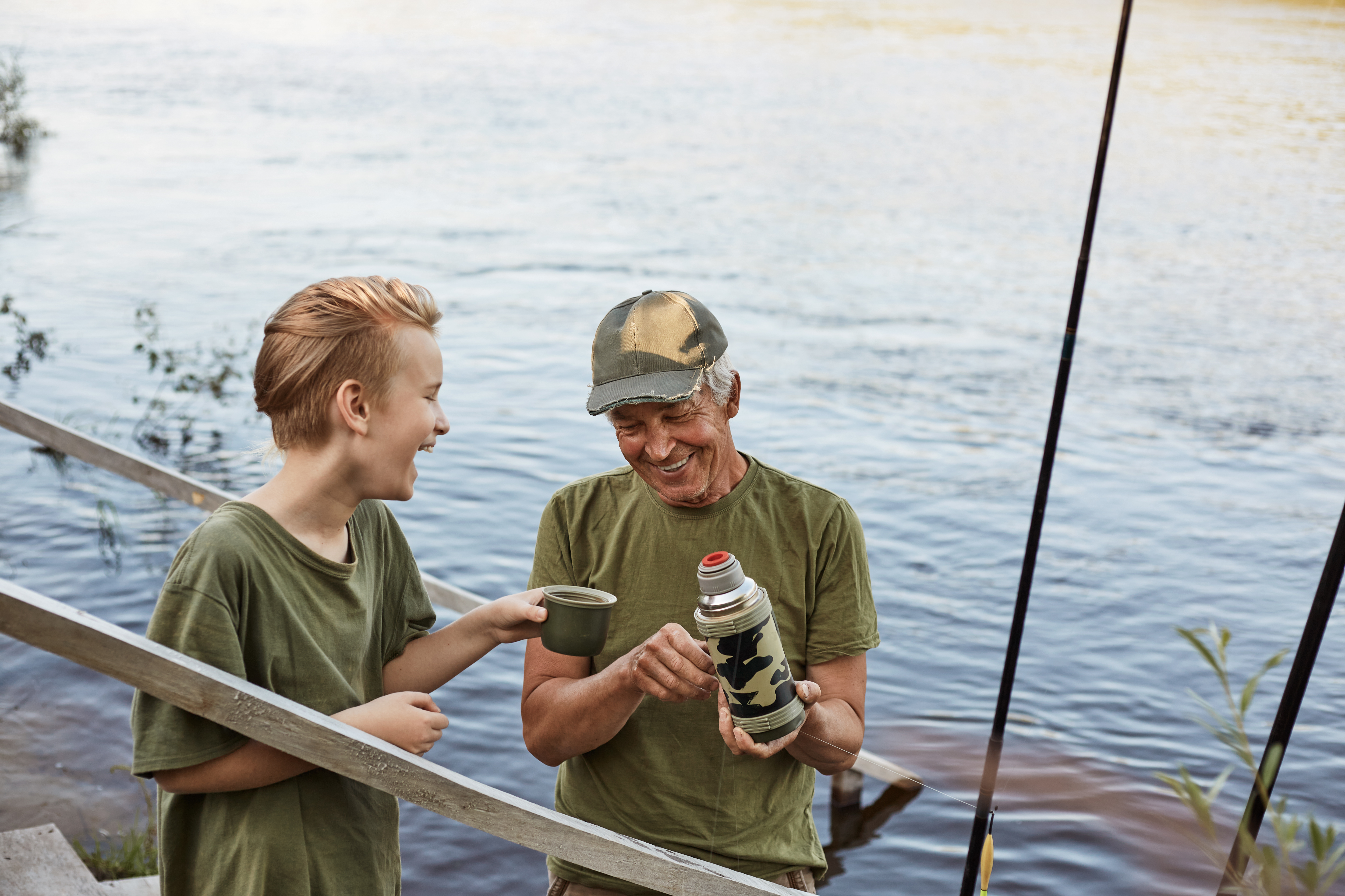 Grandfather and boy fishing together, boy and old man standing with fishing rod, drinking hot beverage from thermos, spending time together and enjoying beautiful nature.