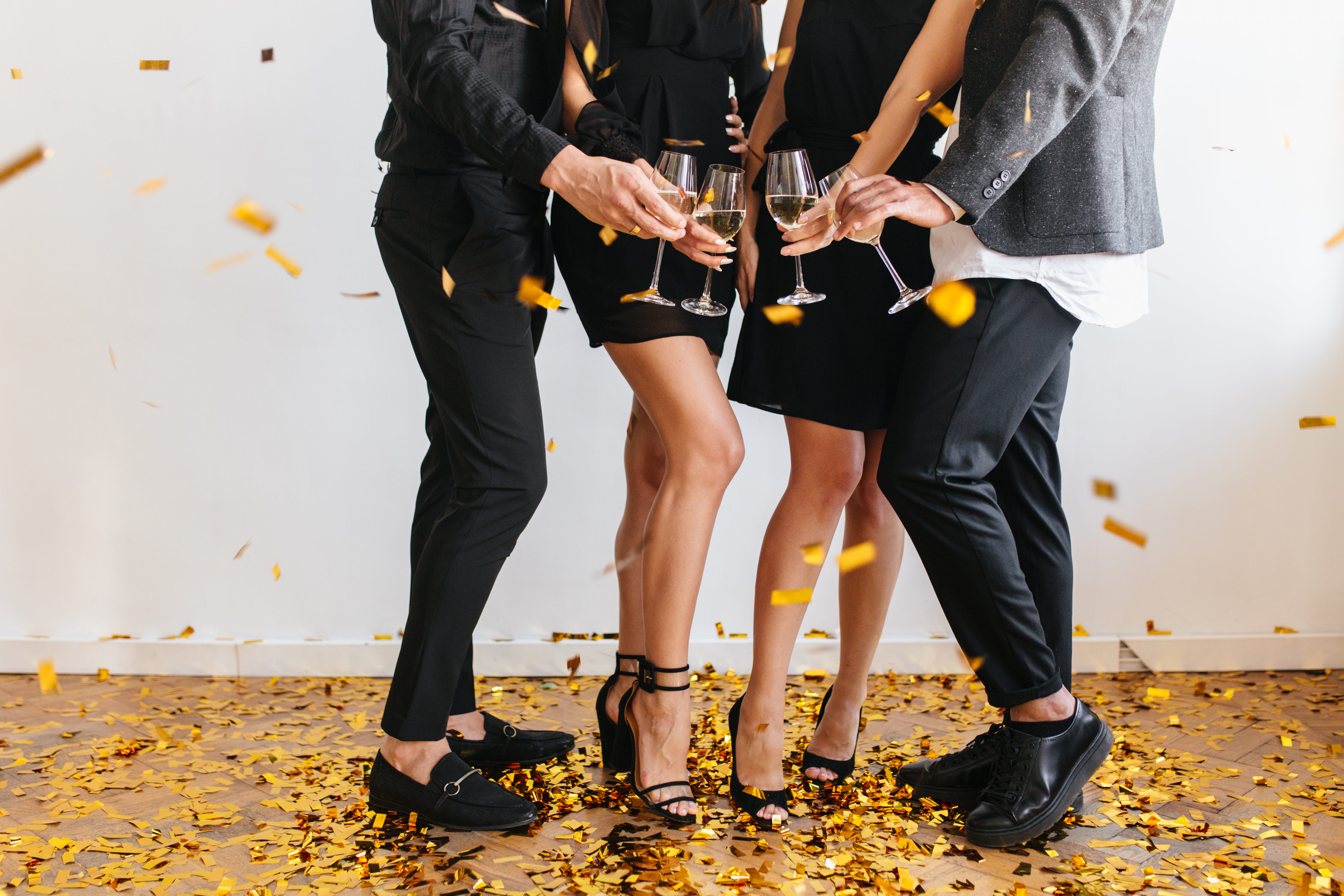 Lightly-tanned lady in trendy sandals standing on the floor covering sparkle confetti. Indoor portrait of boys and girls holding glasses, enjoying cool birthday party together..